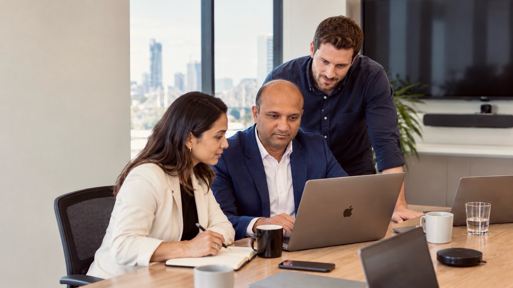 Three members of the TechFluent Systems team working through a build around a laptop, with the Brisbane skyline behind them